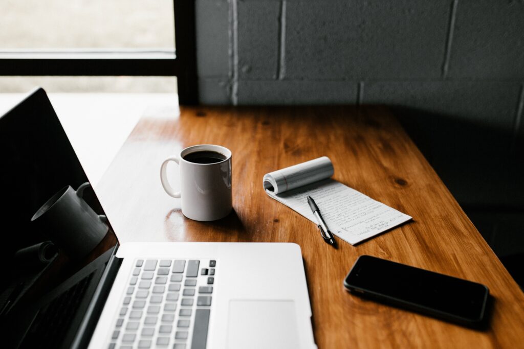 Wooden desk with a laptop next to a cup of coffee, cell phone, pen, and pad of paper